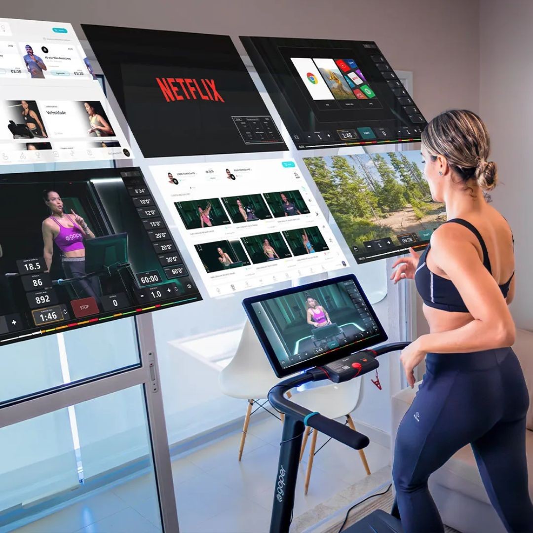 Mulher de costas, com cabelo preso e roupa de gin&aacute;stica preta, correndo em esteira moderna com tela que exibe uma aula. Ao redor, v&aacute;rias telas flutuantes mostram aplicativos de treino, Netflix e paisagens, sugerindo op&ccedil;&otilde;es de entretenimento e exerc&iacute;cios