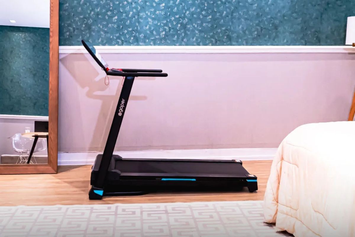 Esteira ergom&eacute;trica preta com detalhes azuis, marca egoper, em um quarto com parede rosa claro e papel de parede azul texturizado. Um espelho de corpo inteiro reflete uma mesa com notebook e cadeira transparente. &Agrave; direita, parte de uma cama com edredom bege. O ch&atilde;o &eacute; de madeira clara com um tapete estampado