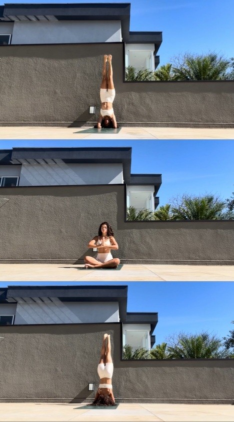 Mulher de cabelo cacheado escuro, top e shorts brancos, praticando yoga em tr&ecirc;s poses: invertida, sentada com m&atilde;os em prece, e invertida novamente, sobre um tapete verde em um terra&ccedil;o com parede cinza e c&eacute;u azul