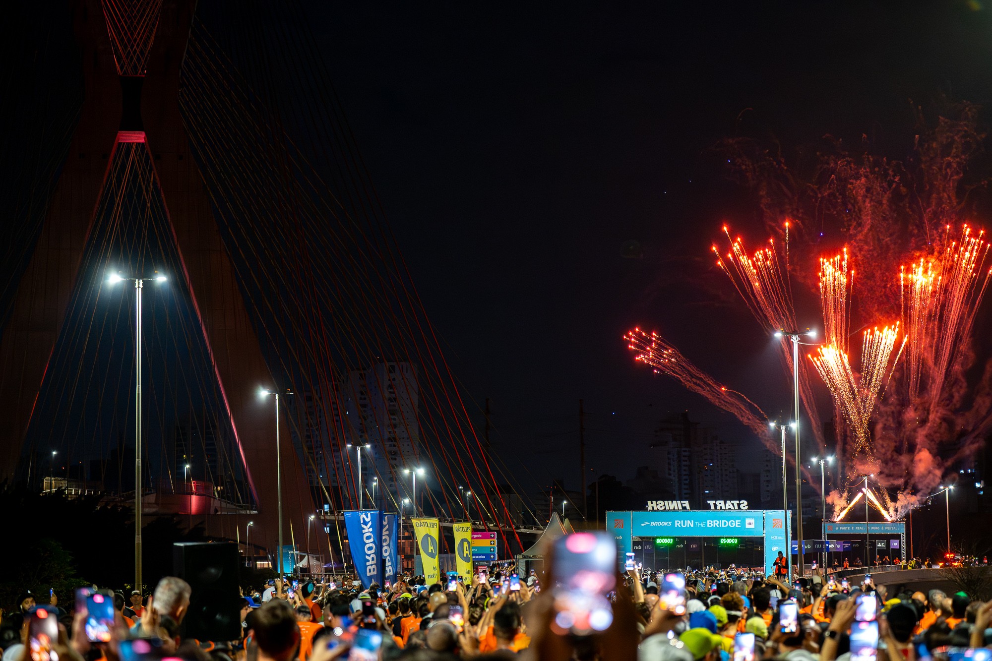 Multid&atilde;o de pessoas com celulares erguidos filmando fogos de artif&iacute;cio vermelhos explodindo sobre a Ponte Estaiada, iluminada em vermelho, durante a noite. &Agrave; frente, um p&oacute;rtico azul com FINISH START RUN THE BRIDGE e banners de patrocinadores. O c&eacute;u est&aacute; escuro, e postes de luz iluminam a cena.