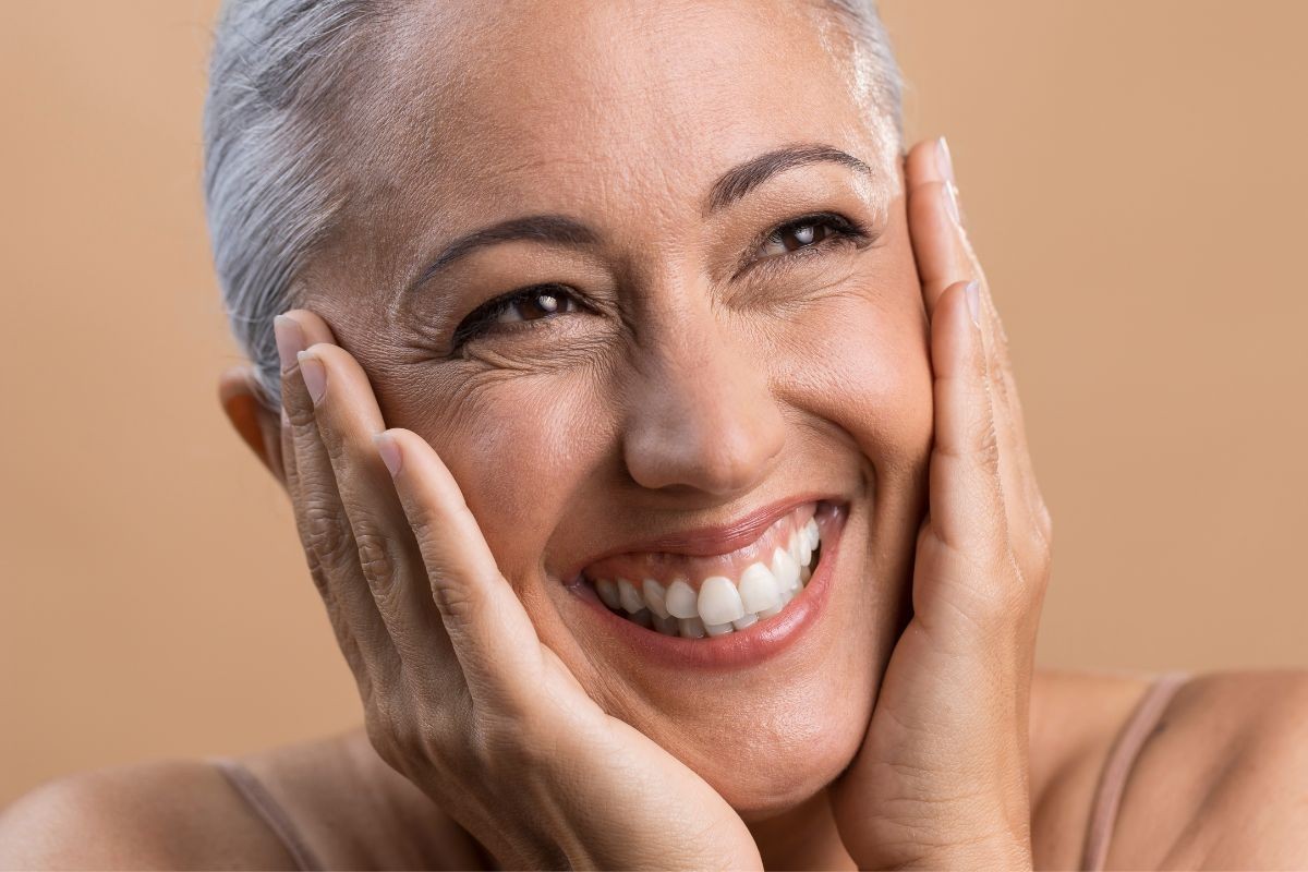 Mulher de pele morena e cabelos grisalhos curtos sorrindo amplamente, com as mãos nas bochechas, sobre fundo bege