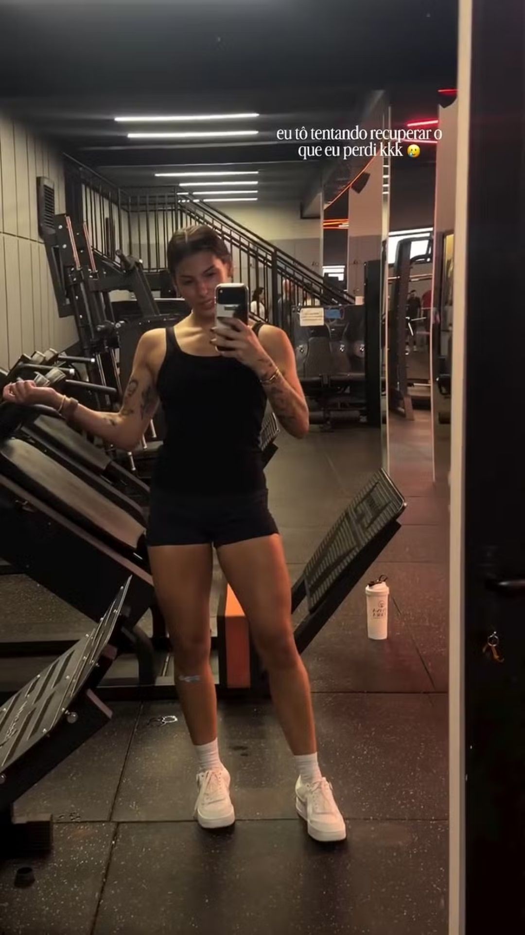 Mulher jovem, morena, com cabelo preso, tira selfie no espelho de uma academia. Ela veste top preto, shorts preto e t&ecirc;nis branco, exibindo bra&ccedil;os tatuados e pernas musculosas. Ao fundo, equipamentos de muscula&ccedil;&atilde;o e uma escada. No canto superior direito, texto: eu t&ocirc; tentando recuperar o que eu perdi kkk. Um copo branco est&aacute; no ch&atilde;o, &agrave; direita
