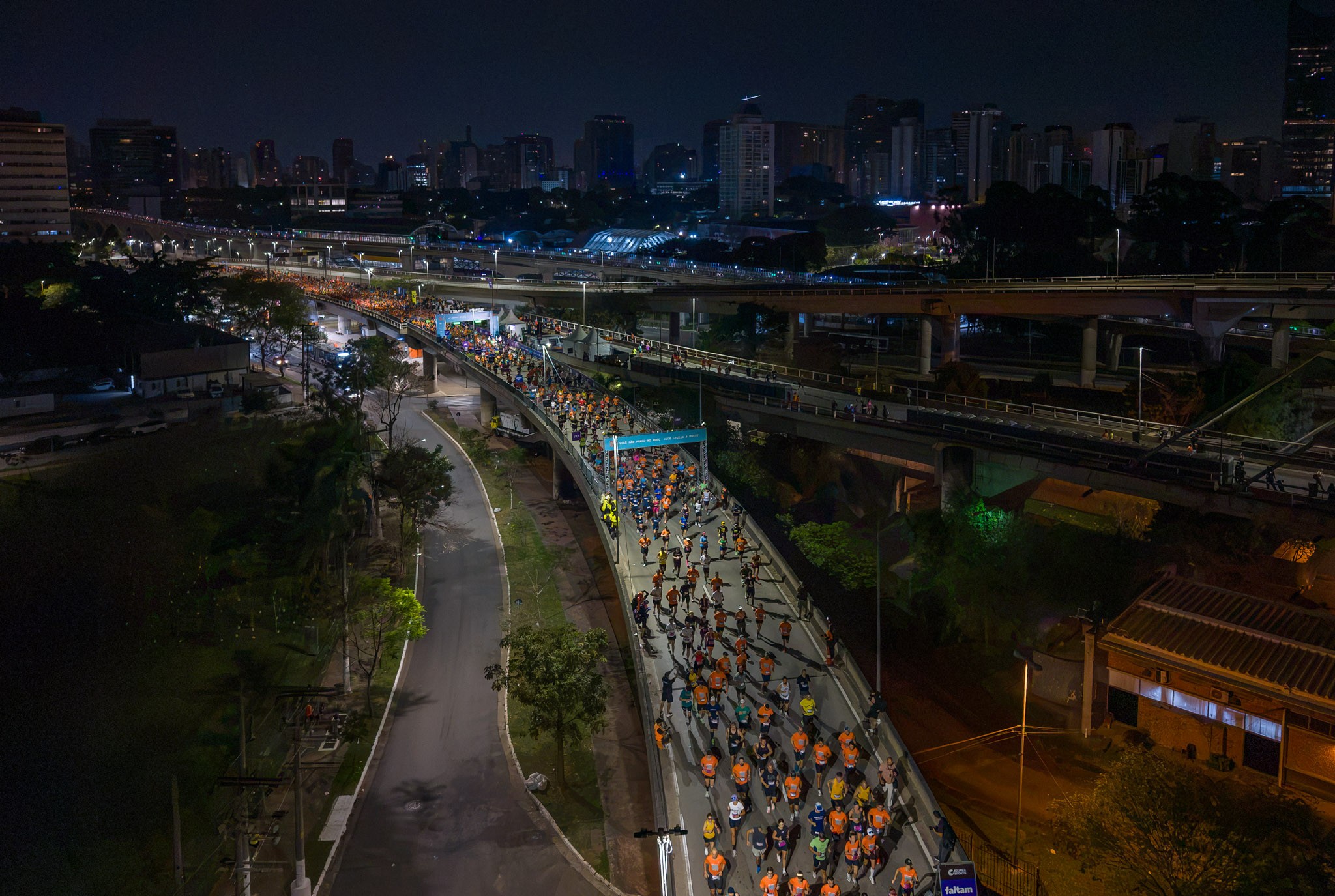 Vista a&eacute;rea noturna de uma corrida de rua com centenas de pessoas em uma via elevada, usando camisetas coloridas, principalmente laranja e azul. Ao fundo, a silhueta iluminada de pr&eacute;dios urbanos e complexos de viadutos, com algumas &aacute;rvores e vegeta&ccedil;&atilde;o nas laterais da pista. O c&eacute;u est&aacute; escuro, indicando a noite.