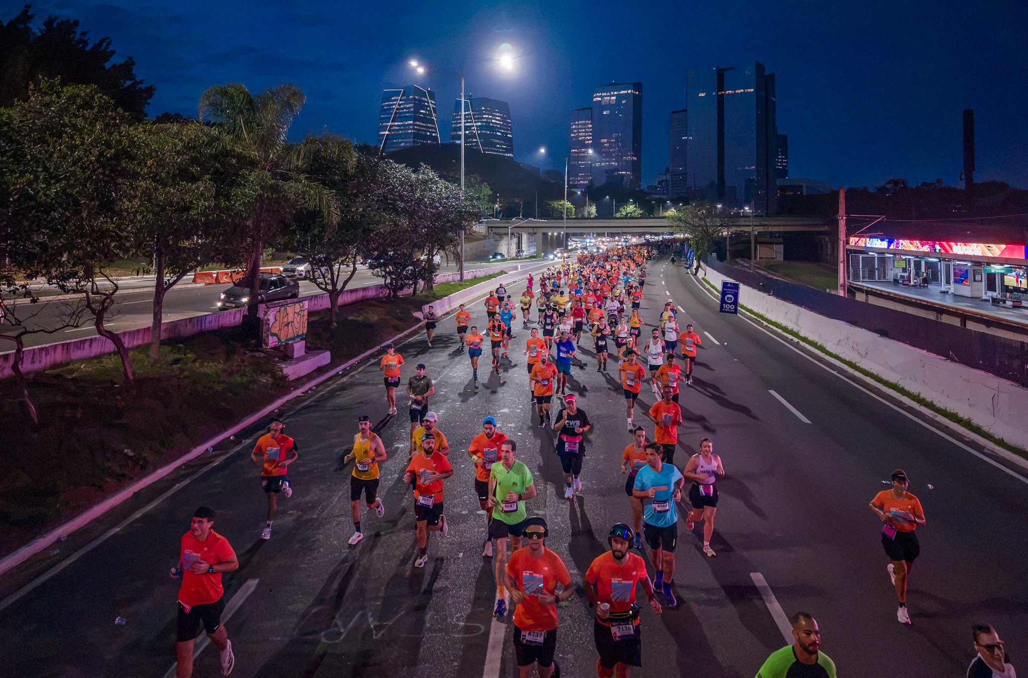 Centenas de corredores participam de uma maratona noturna em uma avenida iluminada, com pr&eacute;dios altos ao fundo e &aacute;rvores nas laterais. A maioria veste camisetas coloridas, principalmente laranja, e shorts, correndo em grupo sob a luz dos postes e da cidade. Um viaduto e uma esta&ccedil;&atilde;o de trem s&atilde;o vis&iacute;veis &agrave; direita