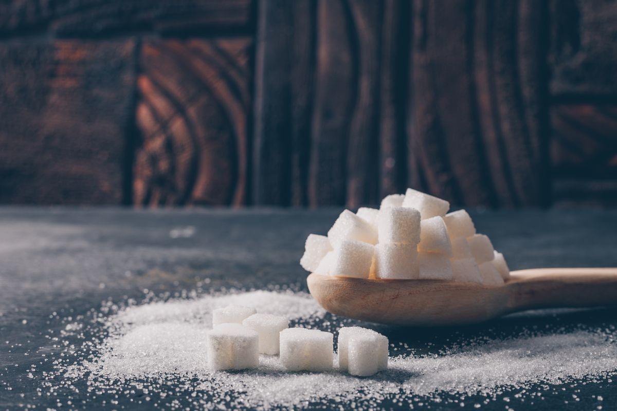 Colher de pau cheia de cubos de açúcar branco, com alguns cubos e açúcar granulado espalhados sobre uma superfície escura, e fundo de madeira marrom