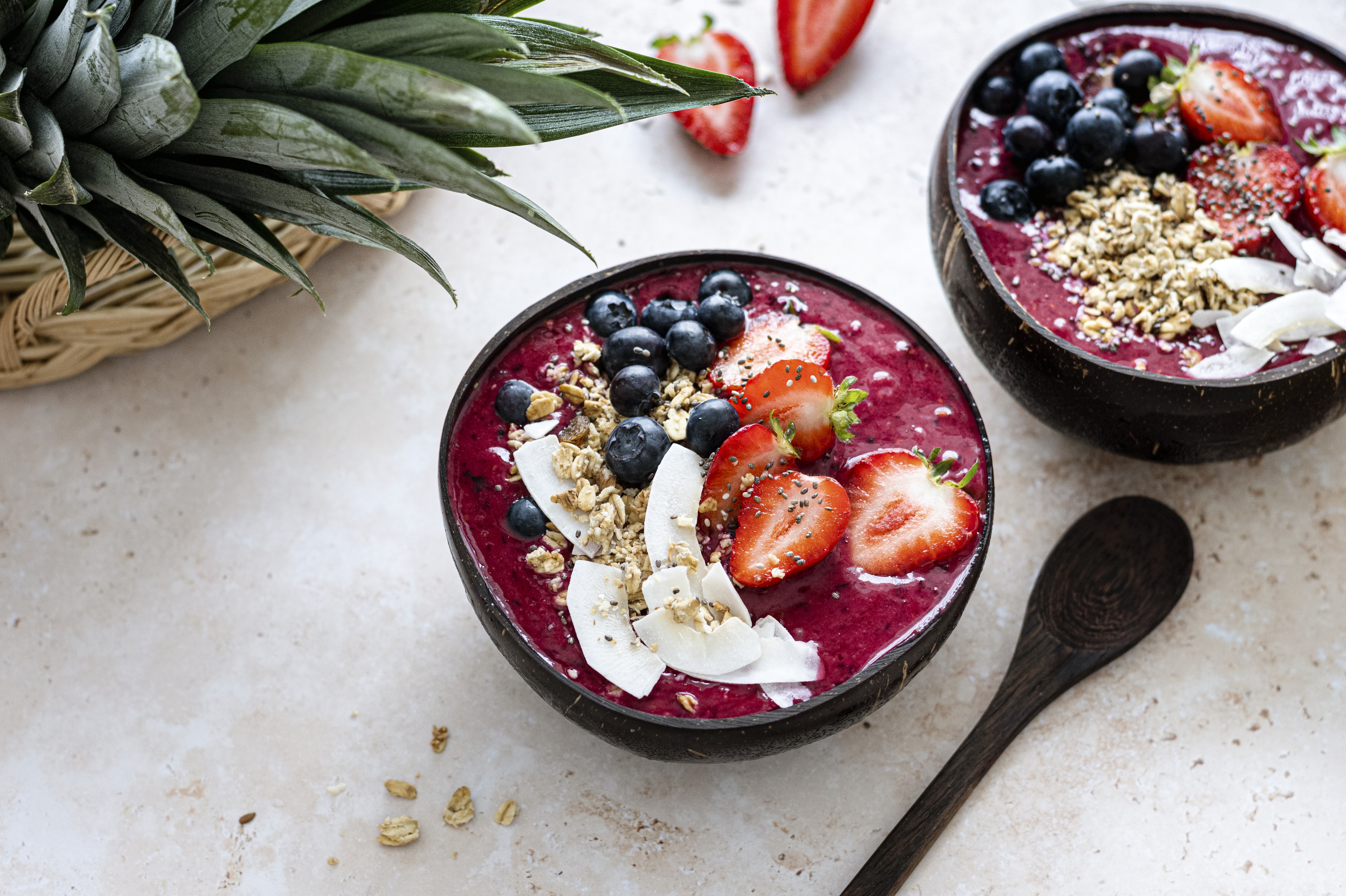Duas tigelas de açaí com mirtilos, morangos fatiados, granola e lascas de coco, sobre uma superfície clara, com um abacaxi e uma colher de madeira ao lado