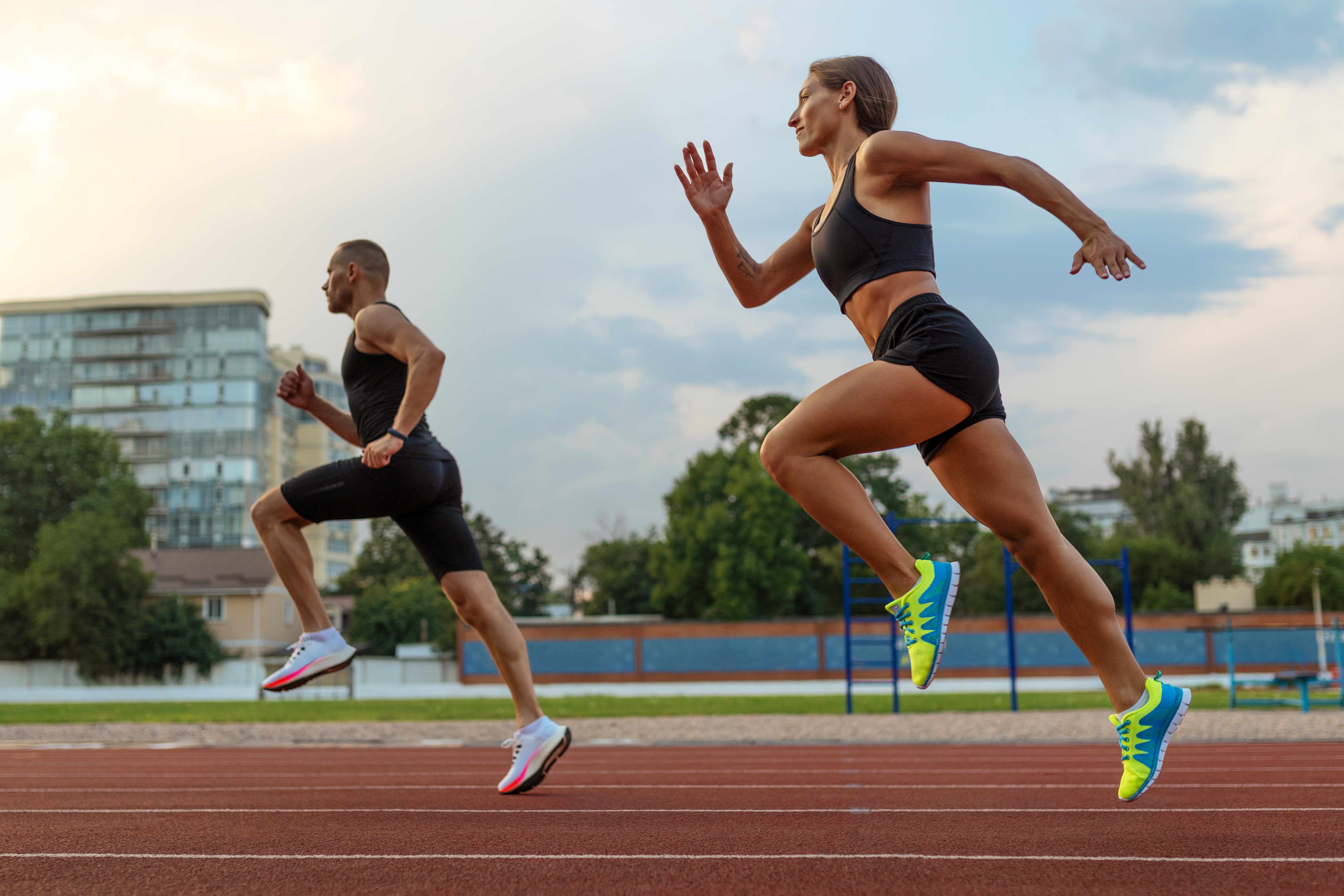 Dois atletas correndo em uma pista de atletismo. A mulher, em primeiro plano, veste top e shorts pretos, tênis azuis e amarelos, com braços flexionados e pernas estendidas. O homem, atrás, usa regata e bermuda pretas, tênis brancos e rosas, com braços e pernas em movimento. Ao fundo, prédios e árvores sob céu nublado